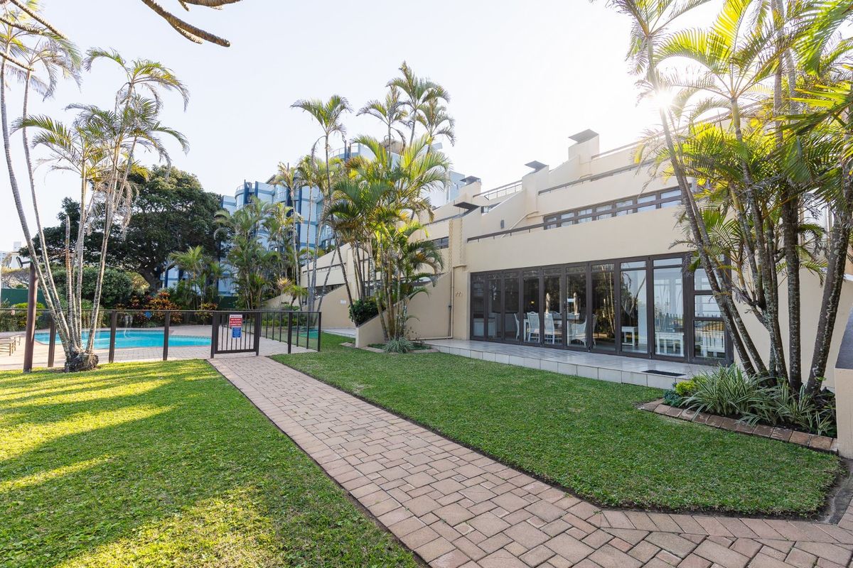 A well-maintained rear exterior view featuring a paved walkway, landscaped garden, and access to a communal swimming pool area with sliding glass doors.