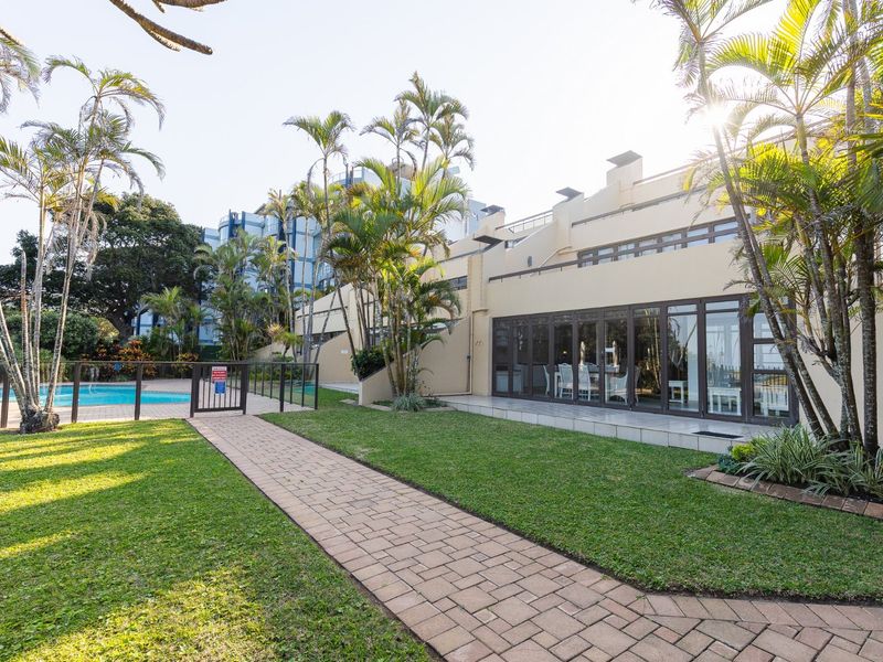 A well-maintained rear exterior view featuring a paved walkway, landscaped garden, and access to a communal swimming pool area with sliding glass doors.
