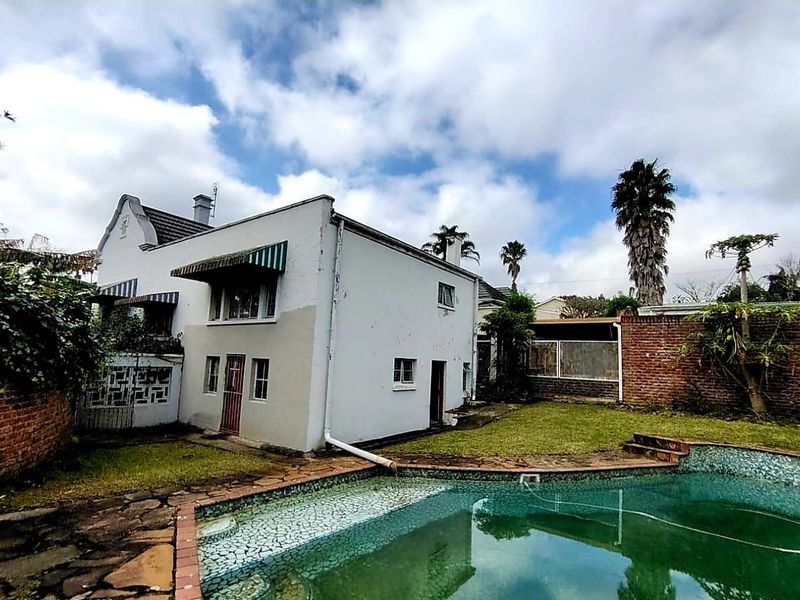 Rear exterior view of the property featuring a swimming pool, brick boundary walls, and a white facade with visible maintenance requirements.