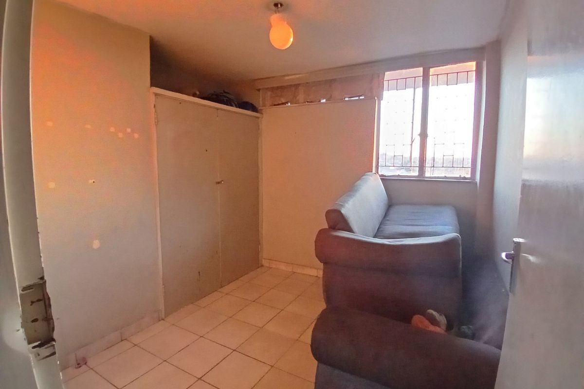 A compact bedroom featuring tiled flooring, a built-in wardrobe, and a window with security bars, requiring cosmetic updates to improve its overall condition.