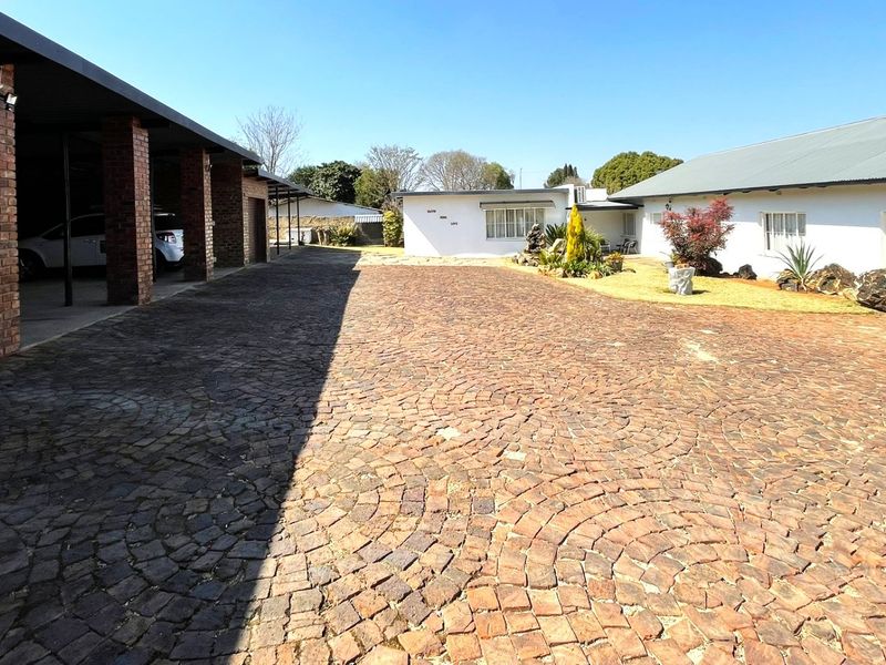 Spacious front exterior featuring a large cobblestone driveway, a brick carport, and well-maintained white building facades under a clear sky.