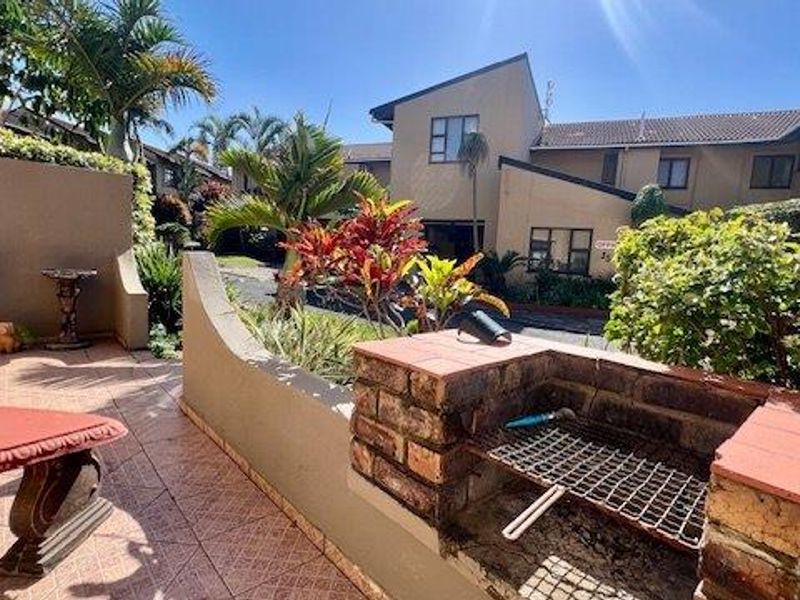 A private outdoor patio area featuring a built-in brick braai, tiled flooring, and established garden surroundings within a townhouse complex.