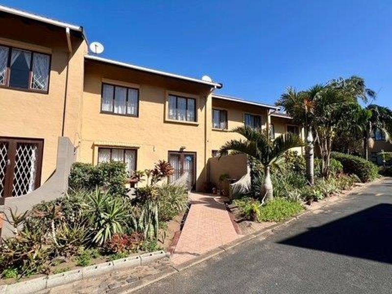 Exterior view of a townhouse complex featuring a paved walkway, established garden beds, and security gates on the ground floor entrances.