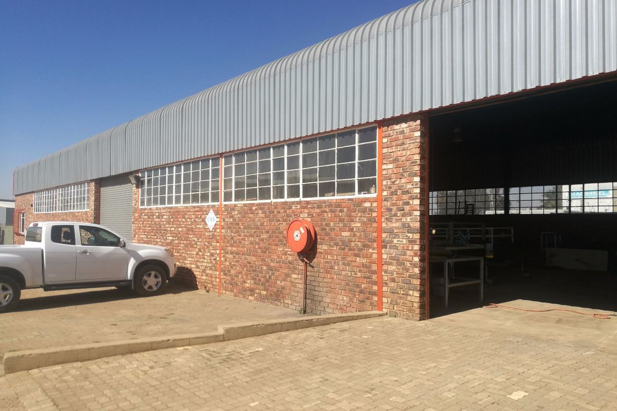 Industrial warehouse facility featuring a face brick facade, corrugated metal roofing, large roller shutter access, and a paved exterior driveway area.