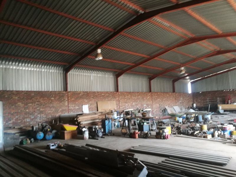 Spacious industrial utility area featuring exposed steel trusses, corrugated metal roofing, brick walls, and concrete flooring, suitable for storage or workshop use.