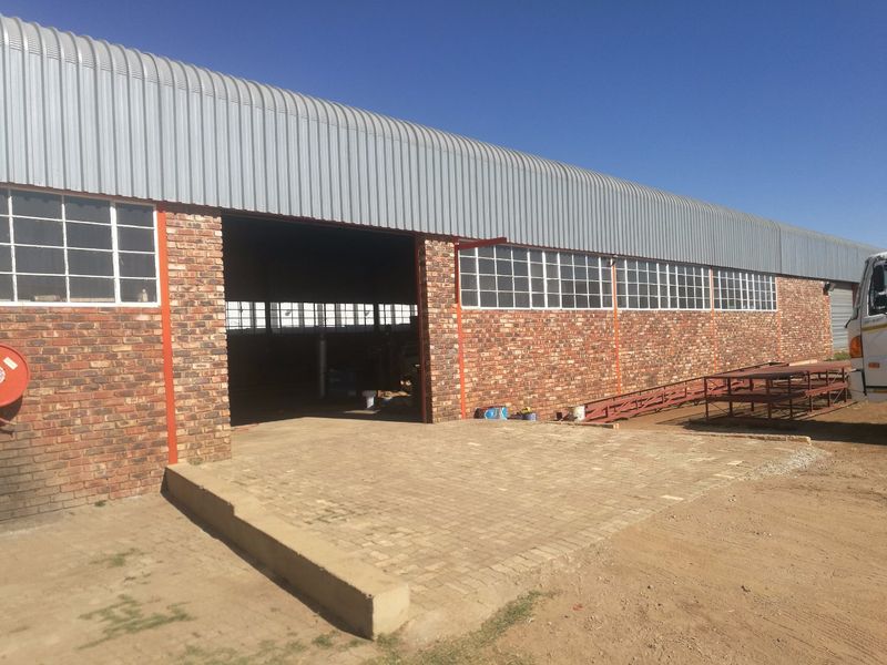 Industrial warehouse exterior featuring a corrugated metal roof, exposed brick facade, large entrance, and a paved loading area.