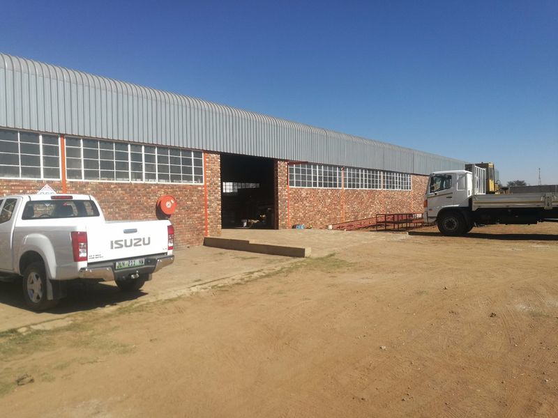 Industrial warehouse facility featuring exposed brickwork, corrugated metal roofing, and large loading bay access, situated on a spacious unpaved yard.