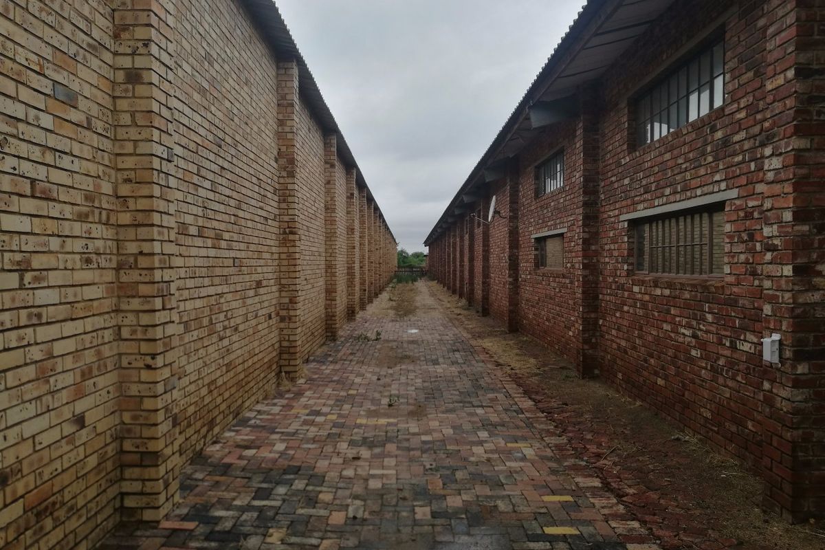 A narrow paved alleyway situated between two industrial brick buildings, featuring exposed brickwork and industrial-style windows.