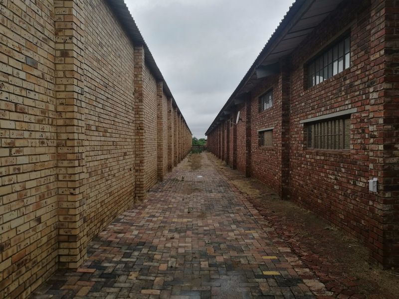 A narrow paved alleyway situated between two industrial brick buildings, featuring exposed brickwork and industrial-style windows.