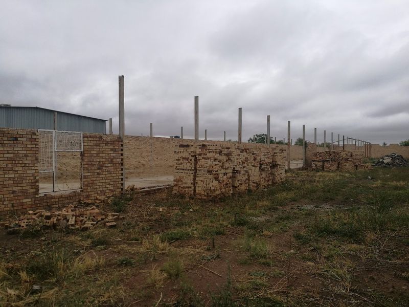 An industrial site under construction featuring a long brick perimeter wall and concrete support columns on an unpaved lot.