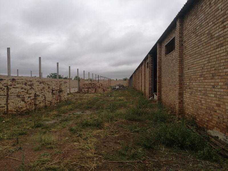 Rear exterior view of an industrial brick building featuring a long facade, concrete boundary posts, and an unpaved yard area.