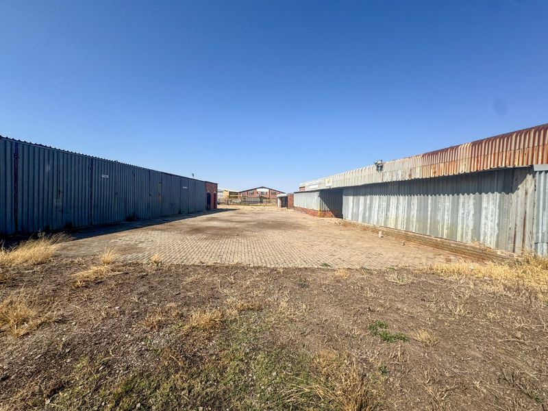 An industrial yard area featuring paved ground and corrugated metal structures on both sides, showing signs of weathering and age.