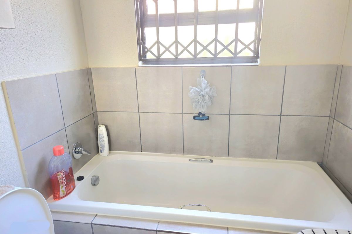 A functional bathroom featuring a built-in bathtub with neutral wall tiling and a window fitted with a security gate.