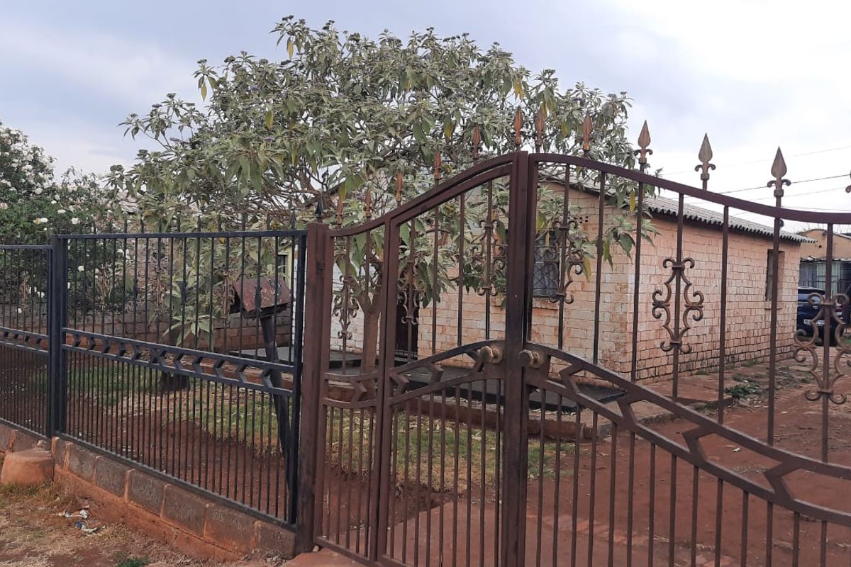 Front view of a single-story brick house featuring a wrought iron gate and perimeter fencing, set on an unpaved plot.