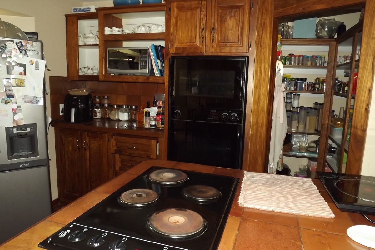 A traditional kitchen featuring extensive wooden cabinetry, a tiled island countertop with an integrated electric hob, built-in oven, and an adjacent walk-in pantry.