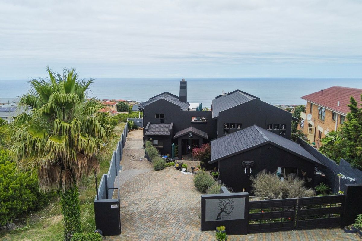 Aerial view of a modern dark-painted house with a paved driveway, perimeter fencing, and ocean views in the background.