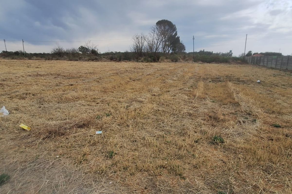 A wide-angle view of a vacant residential plot featuring dry grass, perimeter fencing, and surrounding neighborhood structures.