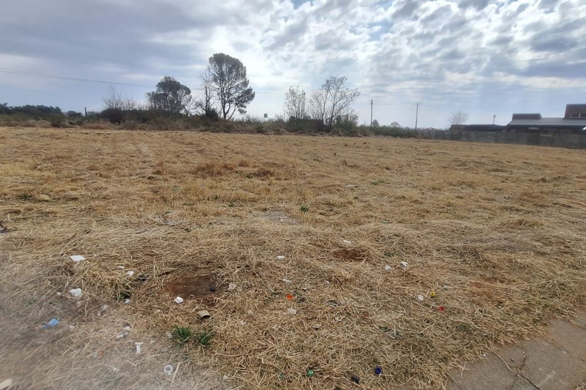 A vacant residential plot of land featuring dry grass, a utility pole, and a boundary wall in the background.