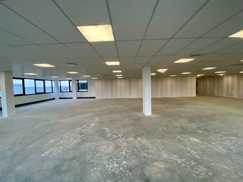 A spacious, open-plan commercial office area featuring a suspended ceiling, recessed lighting, and large windows, currently in an unfinished shell condition.