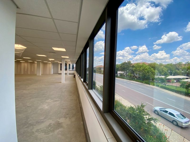 An open-plan commercial office space featuring large windows with street views, suspended ceilings, and unfinished interior surfaces ready for tenant fit-out.