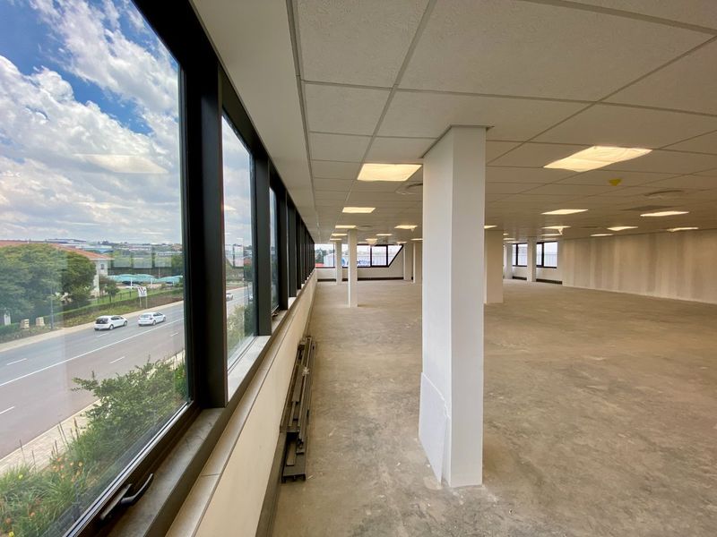 A spacious, open-plan commercial office area featuring large windows with street views, suspended ceilings, and structural columns, currently in a shell condition.