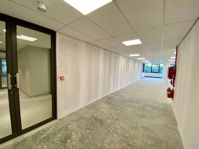 An unfinished commercial hallway featuring a suspended ceiling, recessed lighting, and exposed concrete flooring, currently undergoing renovation with unpainted drywall.