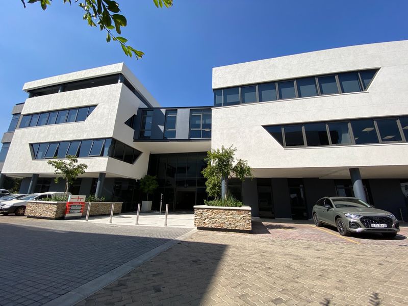 Modern commercial office building featuring a contemporary white facade, extensive glass windows, paved driveway, and integrated stone-clad planters at the main entrance.