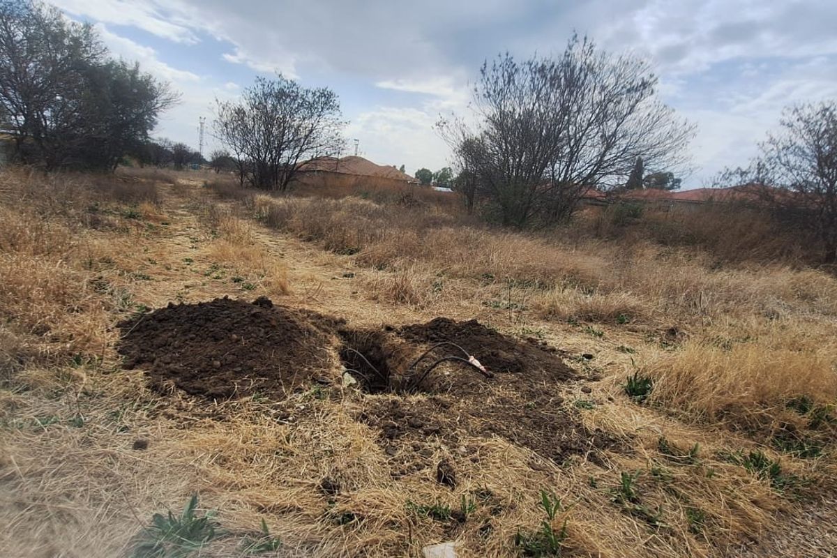 A vacant residential plot featuring dry grass and an open excavation pit with exposed utility cables.
