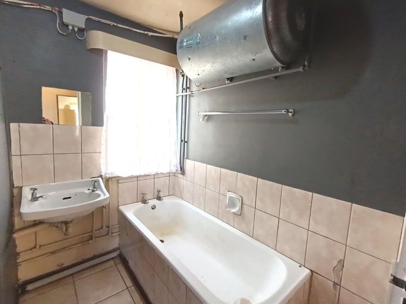 A dated bathroom featuring a bathtub, wall-mounted basin, and exposed water heater, requiring significant renovation and cosmetic updates to improve functionality and aesthetics.