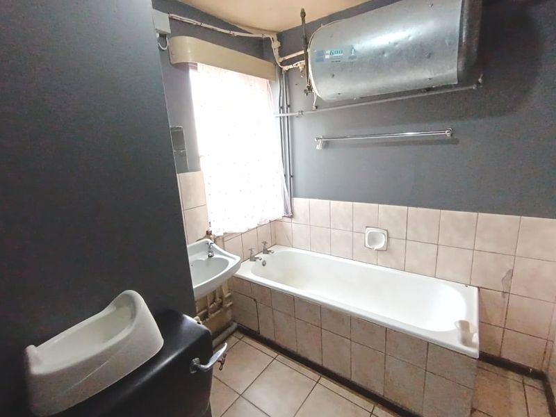 A dated bathroom featuring a bathtub, washbasin, and toilet, with exposed water heater and plumbing, requiring renovation to modernize the space.