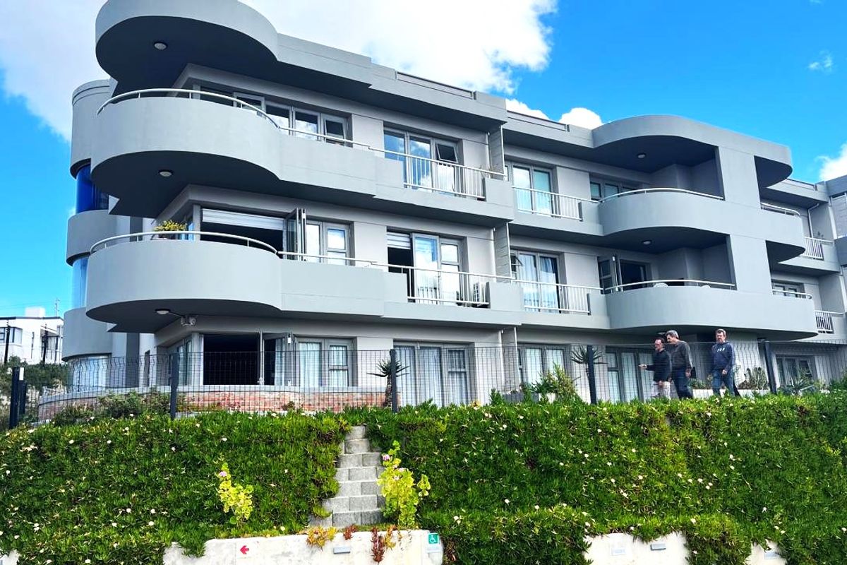 Rear elevation of a modern apartment building featuring curved balconies, metal railings, and sliding glass doors overlooking a landscaped retaining wall.