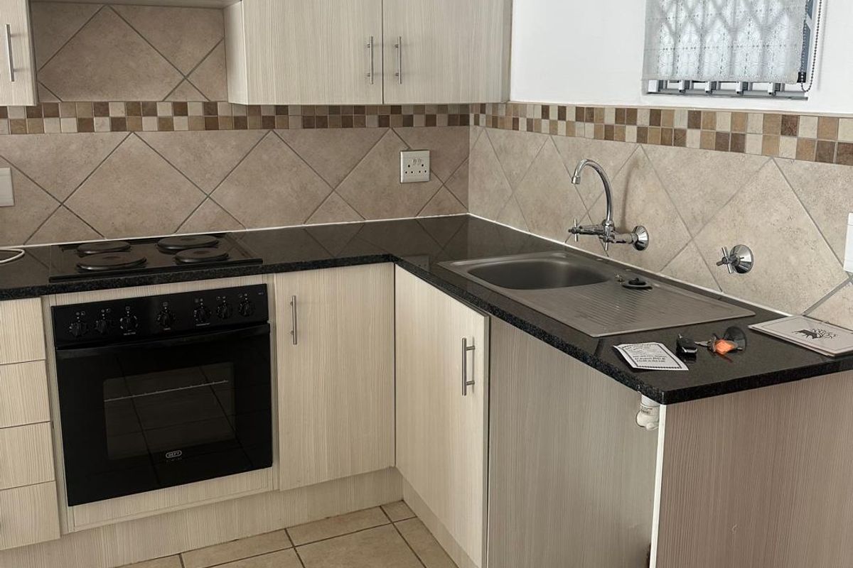 A functional kitchen featuring light wood-grain cabinetry, dark laminate countertops, a built-in electric oven and hob, and a stainless steel sink with tiled backsplash.