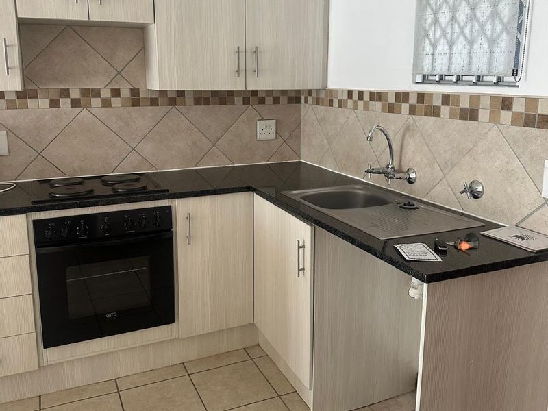 A functional kitchen featuring light wood-grain cabinetry, dark laminate countertops, a built-in electric oven and hob, and a stainless steel sink with tiled backsplash.