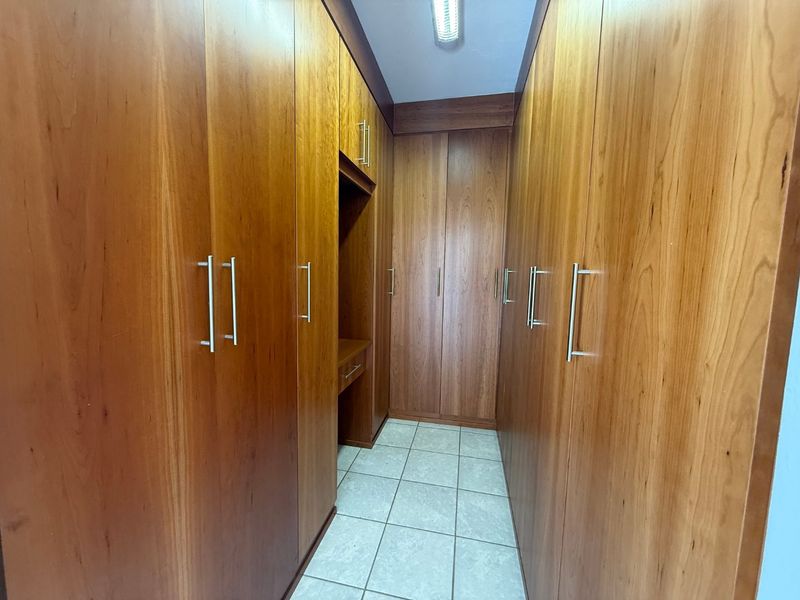 A walk-in closet or dressing area featuring extensive floor-to-ceiling wooden built-in cabinetry, integrated vanity desk, and neutral tiled flooring.