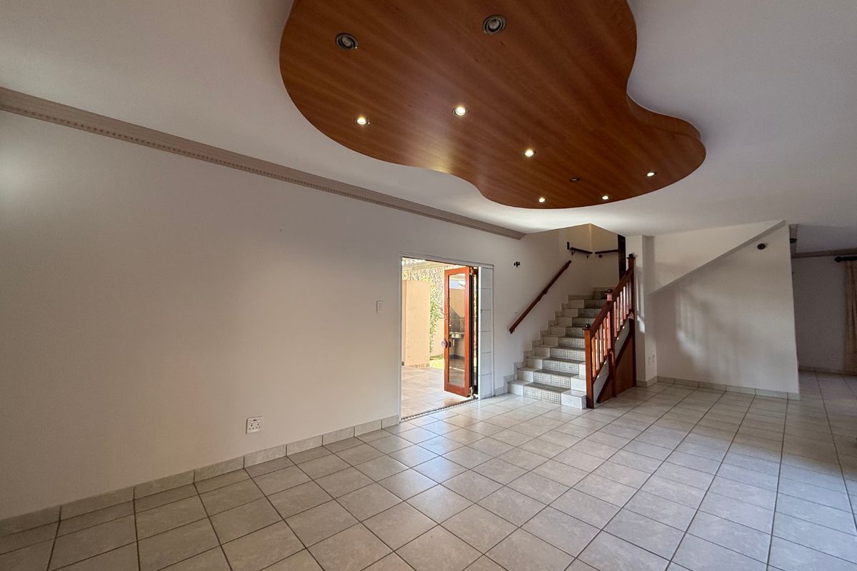 Spacious open-plan living area featuring tiled floors, a unique decorative wooden ceiling with recessed lighting, and a staircase leading to the upper level.