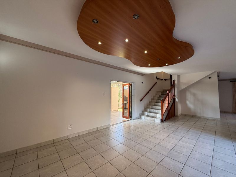 Spacious open-plan living area featuring tiled floors, a unique decorative wooden ceiling with recessed lighting, and a staircase leading to the upper level.