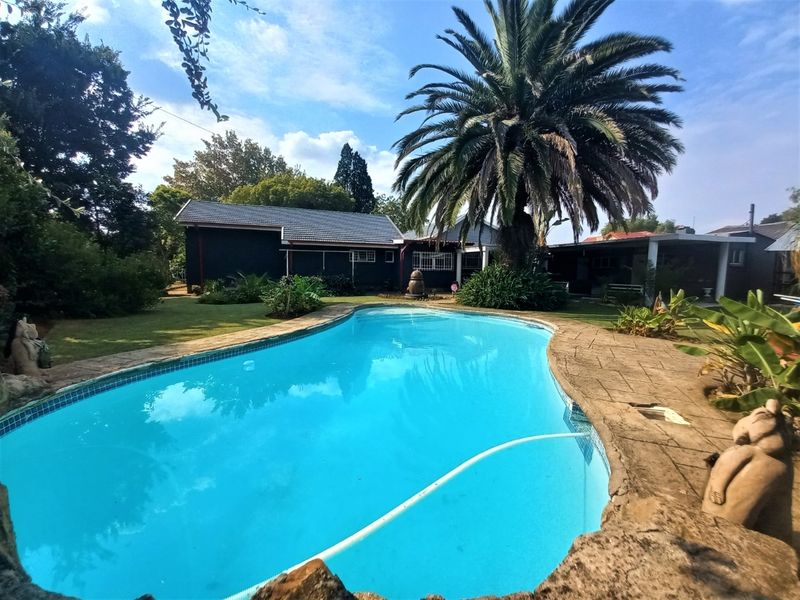 A spacious backyard featuring a large swimming pool with a paved surround, set within a well-established garden with mature trees and a tiled-roof dwelling.