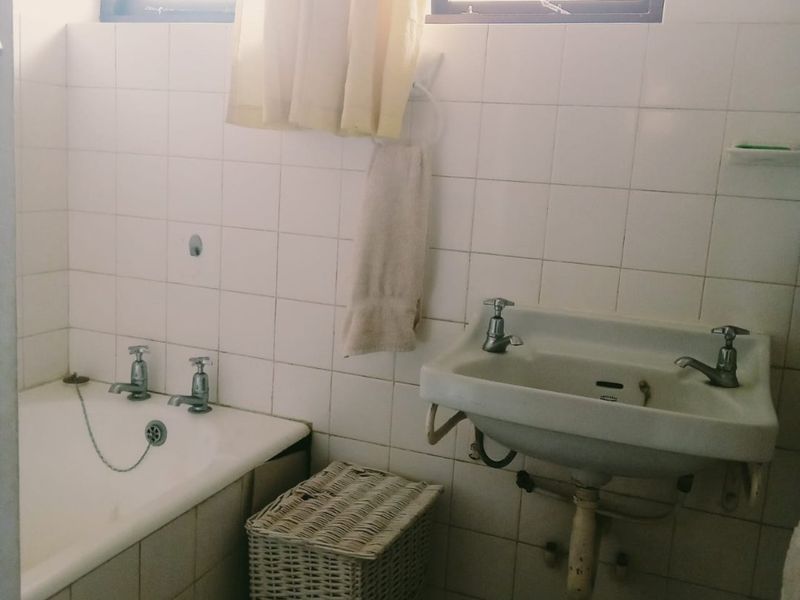 A dated bathroom featuring a wall-mounted basin and a built-in bathtub with original chrome fixtures and white ceramic wall tiling.