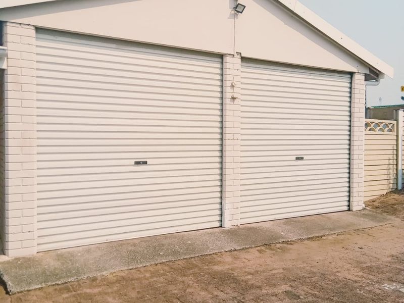 A double garage featuring white roller doors and a brick facade, set against a concrete apron and adjacent boundary wall.