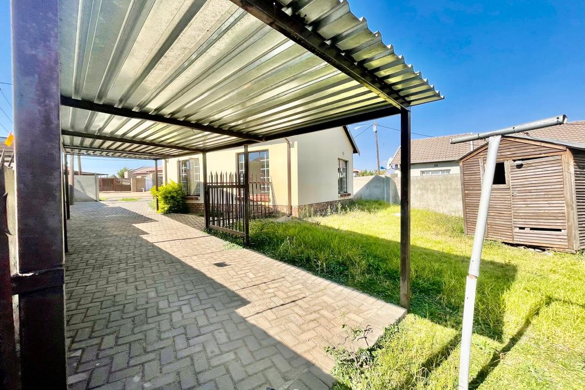 Rear exterior view featuring a paved driveway with a corrugated metal carport, a wooden garden shed, and a perimeter wall.