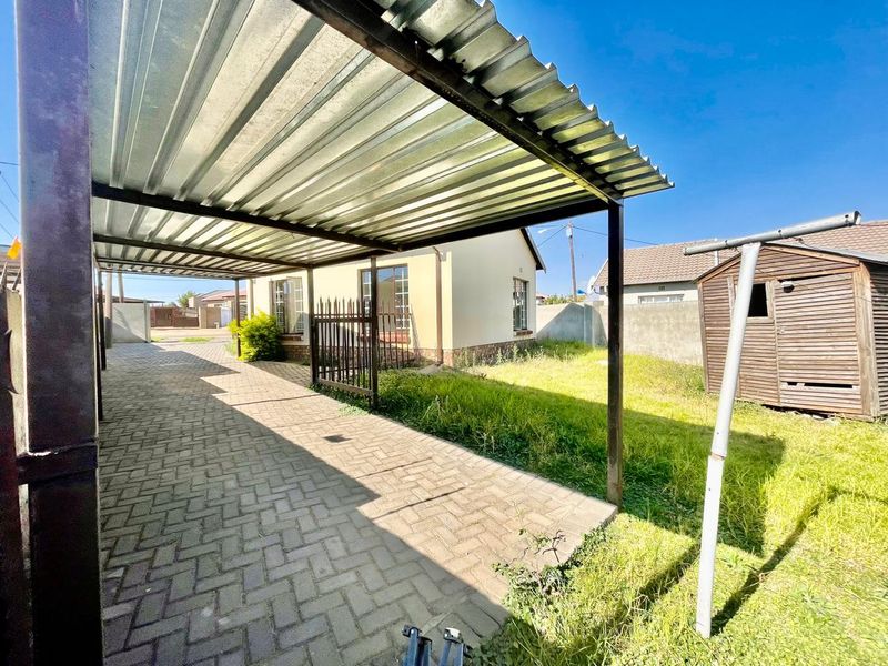 Rear exterior view featuring a paved driveway with a corrugated metal carport, a wooden garden shed, and a perimeter wall.
