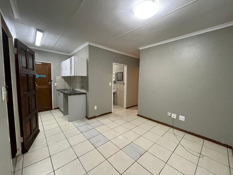 An open-plan living area featuring a compact kitchenette with built-in cabinetry and sink, tiled flooring, and neutral wall finishes.