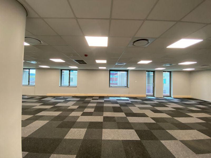 A spacious, open-plan commercial office area featuring modern carpet tiles, a suspended ceiling with recessed LED lighting, and large windows providing natural light.