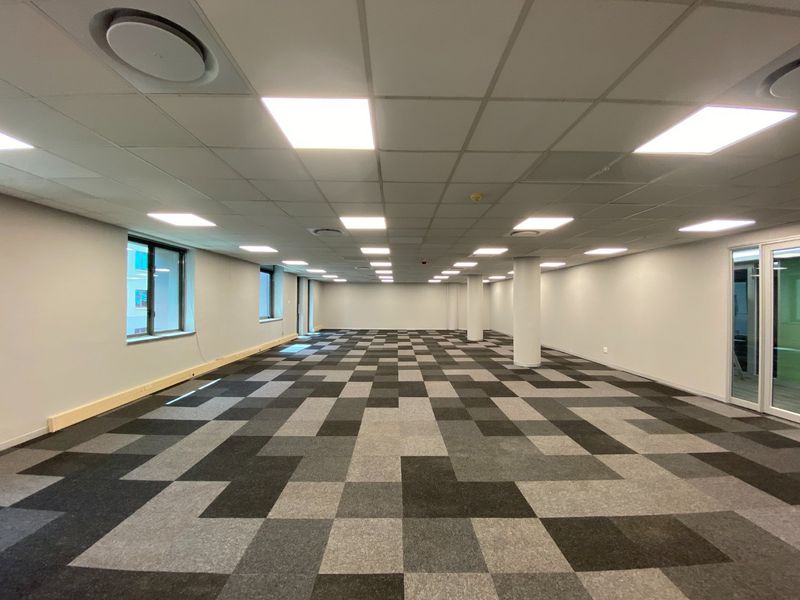 A spacious, open-plan commercial office area featuring a suspended ceiling with recessed lighting, carpet tile flooring, and large windows providing natural light.
