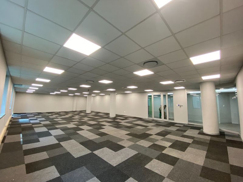 Spacious open-plan commercial office area featuring modern carpet tiles, suspended ceilings with recessed lighting, and glass partition doors.
