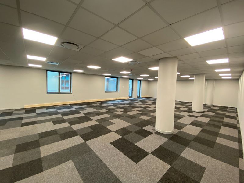A spacious, open-plan commercial office area featuring modern carpet tiles, recessed ceiling lighting, and structural columns, providing a clean and professional workspace.