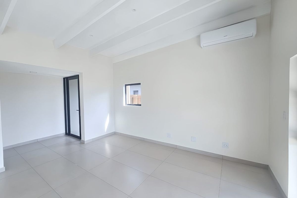 A bright, modern, and empty living area featuring tiled floors, exposed ceiling beams, and a wall-mounted air conditioning unit.