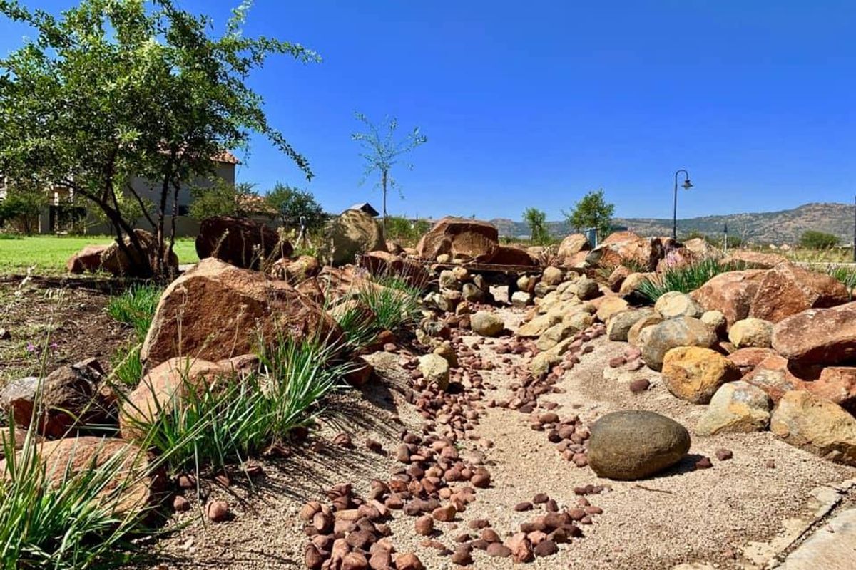 A well-maintained landscaped garden featuring a decorative dry creek bed, natural rock formations, and established greenery under a clear blue sky.