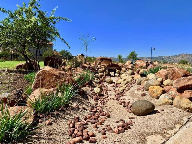 A well-maintained landscaped garden featuring a decorative dry creek bed, natural rock formations, and established greenery under a clear blue sky.
