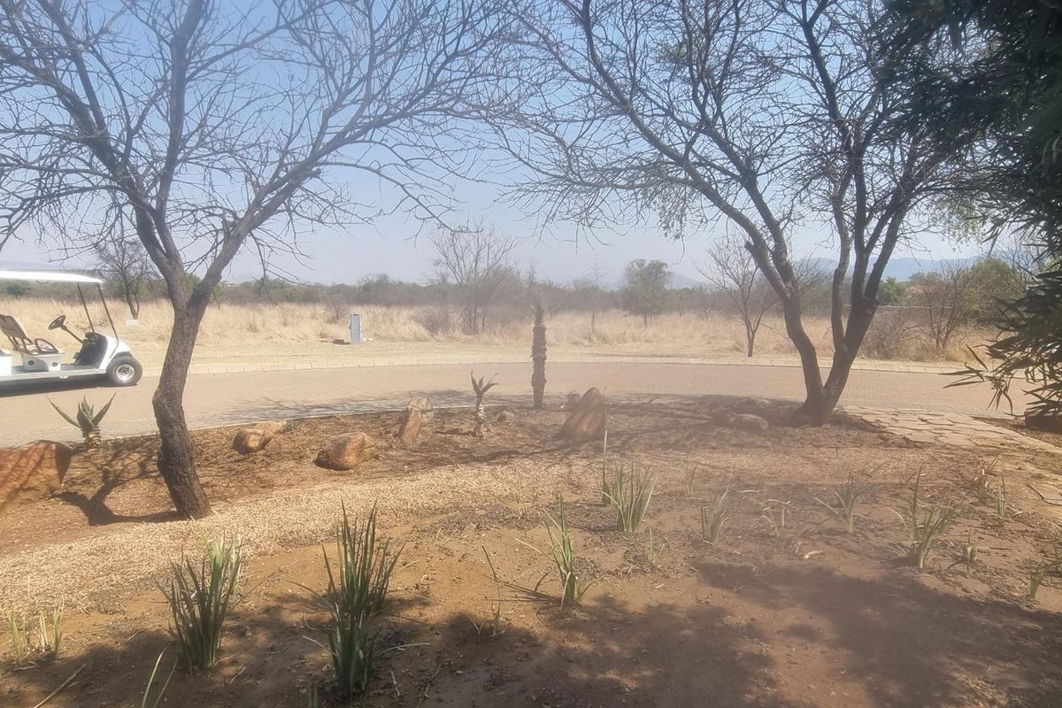 A view of a landscaped garden area featuring mature trees, paved driveway access, and established garden beds on a residential vacant land plot.